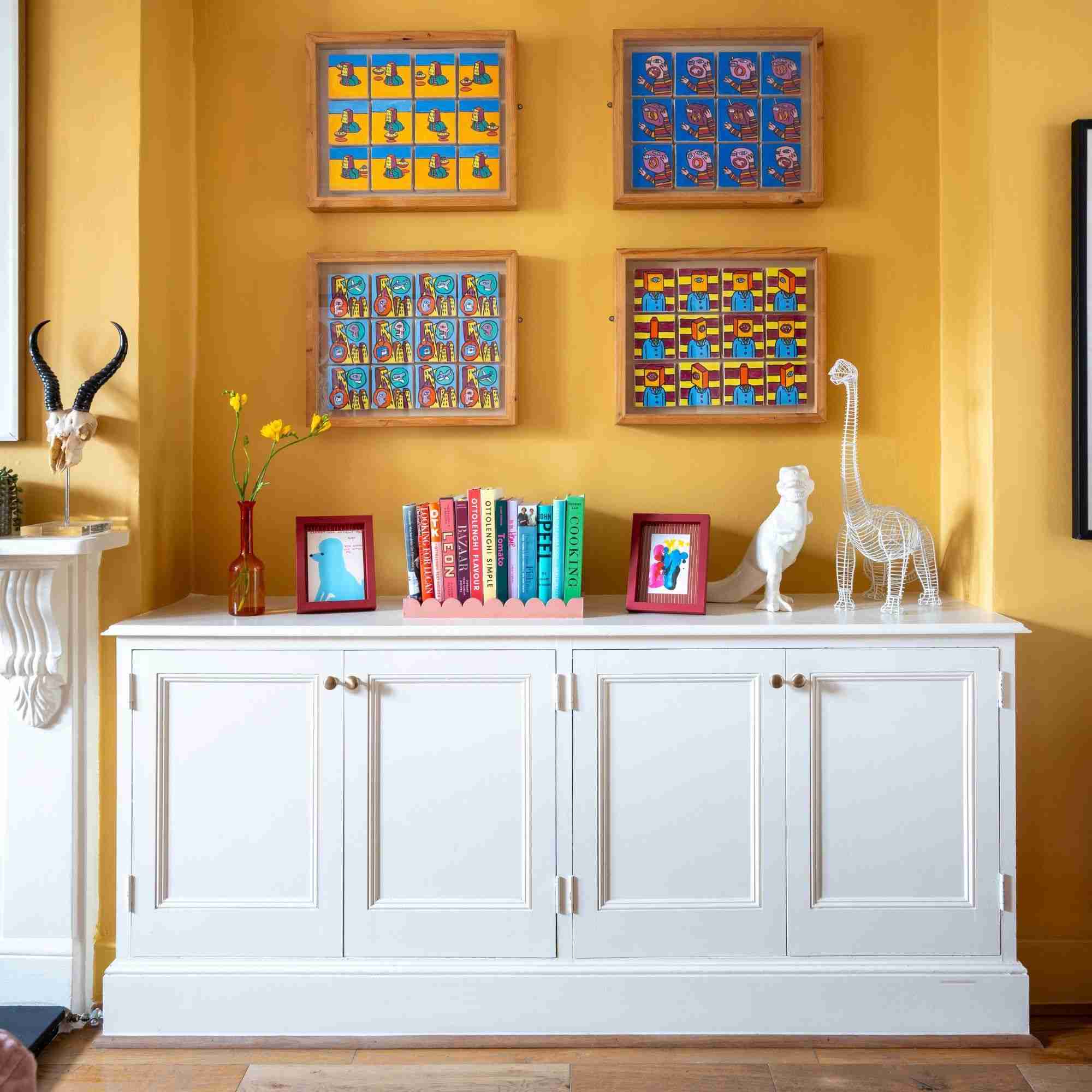 White painted wooden cabinet covered in books and trinkets in front of a sunset yellow wall with artwork on it