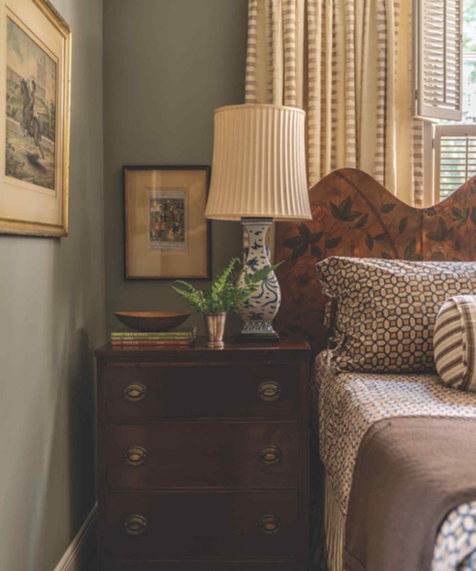 Bedroom with pale blue walls, a mahogany nightstand, a screen headboard, vintage china lamp, patterned geometric bedding, and antique prints on the walls.