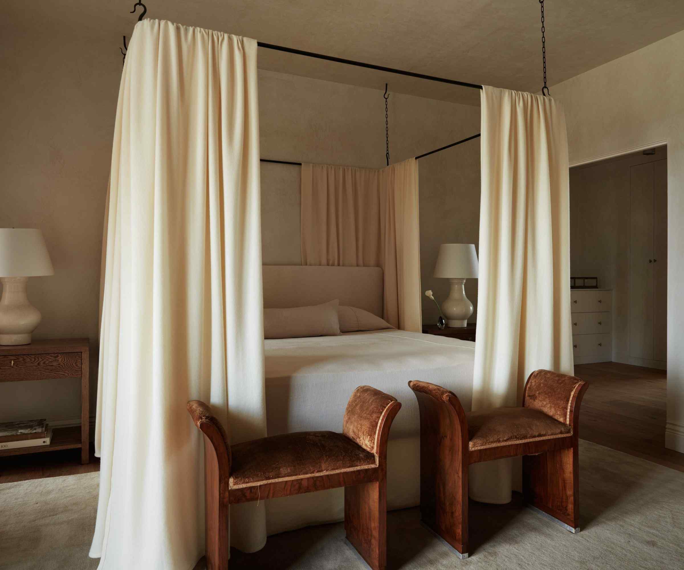 a warm beige bedroom with a four poster bed, drapes, and two small vintage ottomans at the foot of the bed
