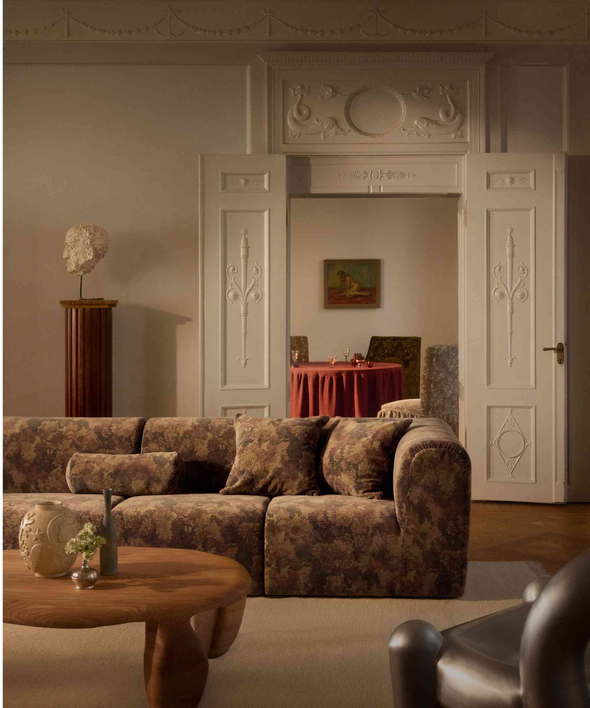 Tapestry-patterned sofa and matching throw pillows styled in an opulent living room with ornate moulding, a Grecian bust, and low-slung, organically-shaped coffee table.