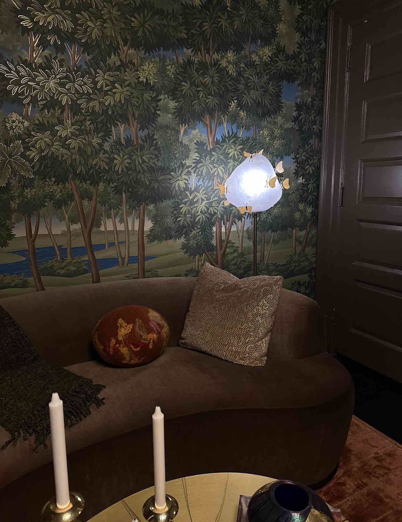 Patterned 'ball' pillow styled atop of a curved mauve sofa inside of a living room that features a nature-inspired wall mural backdrop