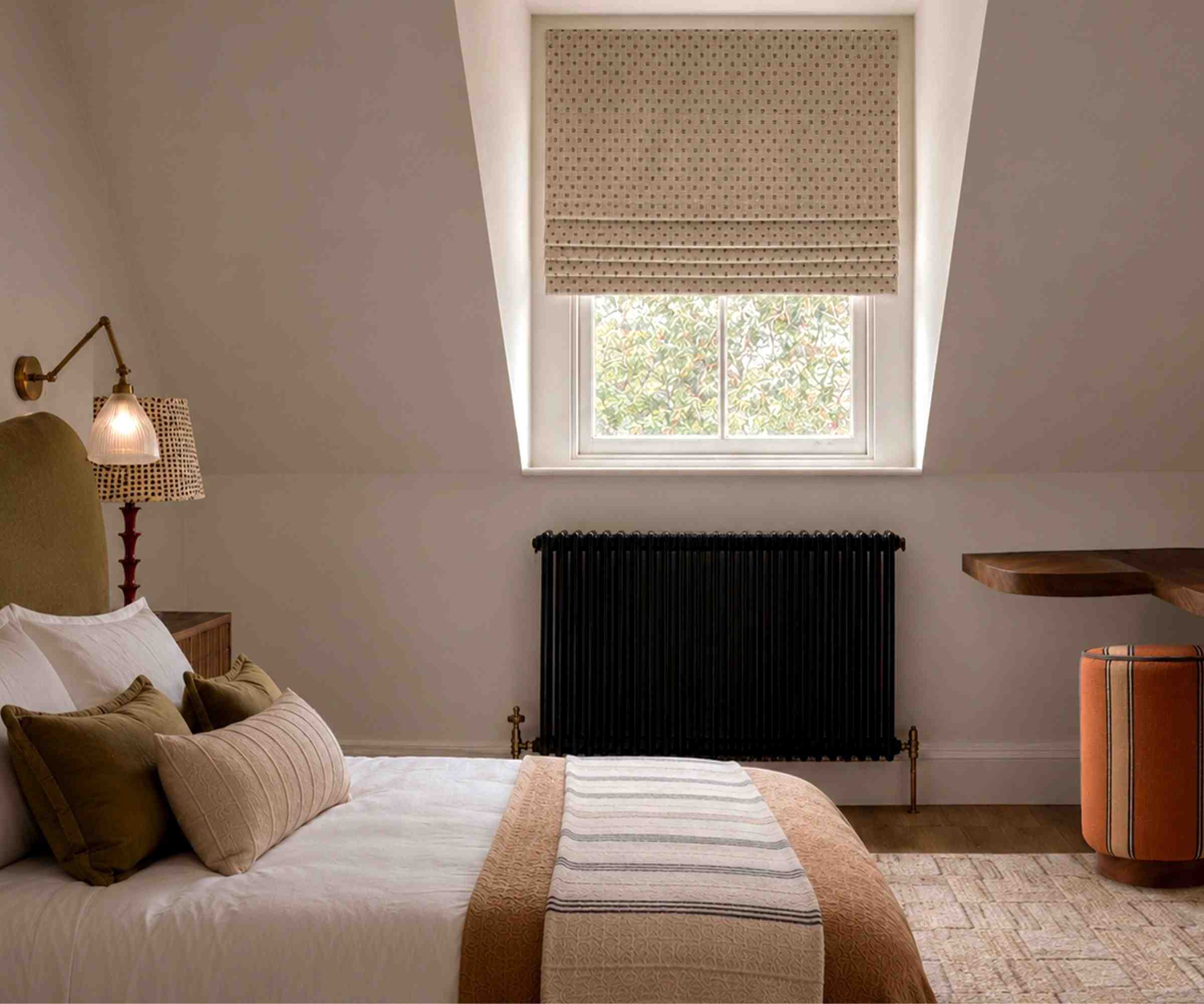 Autumnal bedroom with white walls, a large window with a Roman blind, an olive green bed with neutral bedding and a wool brown throw, a cream rug, with a wall sconce above, and an orange pouffe.