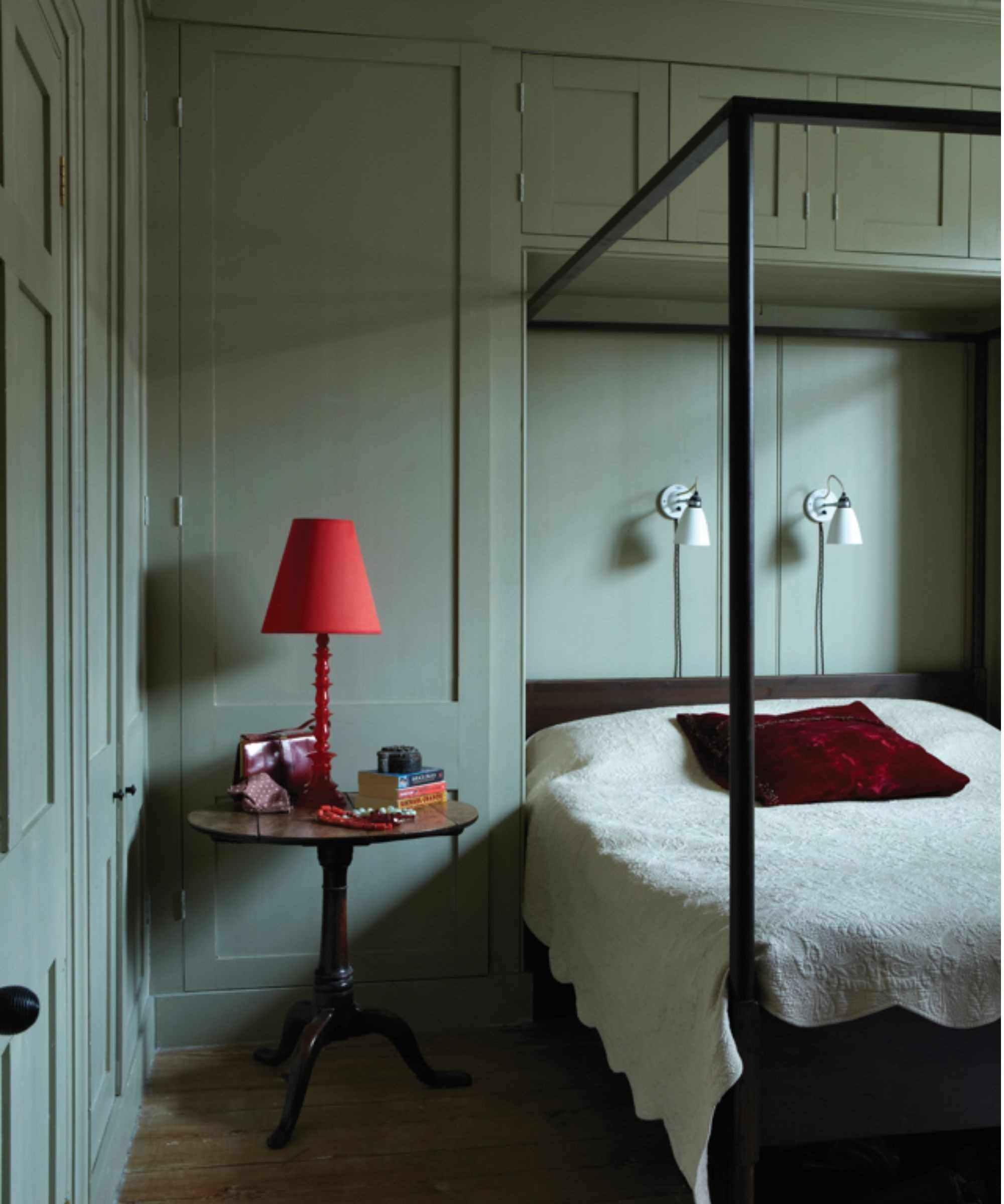 bedroom with color-drenched gray-green walls, a four poster bed, and bright red lamp on a side table